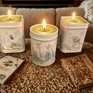 May include: Three lit candles in decorative containers with illustrations. The candles have a square or round shape and are filled with green wax. The containers feature illustrations of animals and garden scenes. A book and a decorative item are also present.
