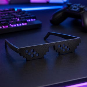 May include: Black pixelated sunglasses featuring a distinct blocky, 8-bit design. The frames are constructed with a matte black finish and square-shaped cutouts that form the lenses, creating a retro digital aesthetic reminiscent of classic video games.