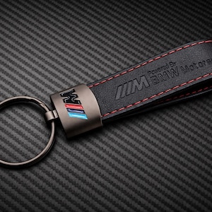 May include: Black leather keychain with red stitching and a metal accent featuring the BMW M logo in blue, red, and white. The keychain has a metal ring and the text "Powered by BMW Motorsport".