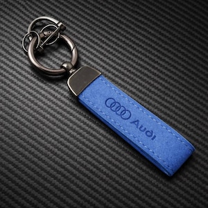 Puede incluir: Llavero Audi de ante azul con cierre y anilla de metal plateado. El logotipo y el texto de Audi son visibles en la correa azul. Fondo texturizado gris oscuro.