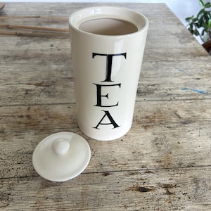 May include: A cylindrical cream-coloured ceramic tea canister with a matching removable lid. The word TEA is printed vertically in large black serif capital letters on the front of the canister. The interior of the canister is smooth and light.