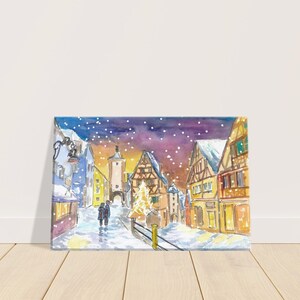 Op de afbeelding: Een aquarel schilderij van een besneeuwd Europees dorp 's nachts. De scène toont een hoge toren, verschillende huizen met besneeuwde daken en een kleine kerstboom in het midden. Twee figuren lopen over straat en de lucht is gevuld met vallende sneeuw.