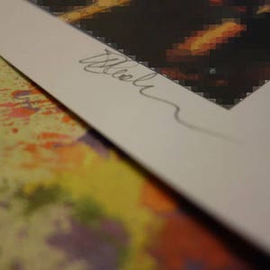 May include: A close-up of a white mat with a handwritten signature in black ink. The mat is placed on top of a colorful, abstract background.