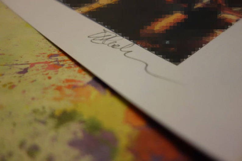 May include: A close-up of a white mat with a handwritten signature in black ink. The mat is placed on top of a colorful, abstract background.