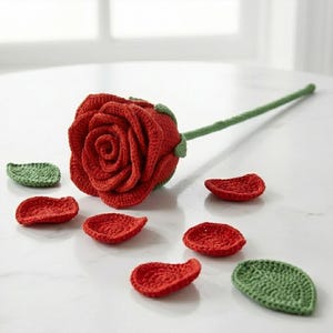 May include: A handcrafted red crochet rose with a green stem and leaves. The rose is surrounded by red crochet petals and green leaves, all arranged on a white surface. The rose is a detailed, handmade craft item.