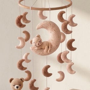 May include: A handmade baby mobile featuring a crocheted bear sleeping on a pink crescent moon, surrounded by smaller crescent moons in varying shades of brown and pink. A crocheted teddy bear sits below.