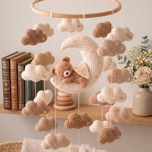 Puede incluir: Móvil para bebé hecho a mano con un oso de ganchillo durmiendo sobre una luna creciente, rodeado de nubes de ganchillo crema y marrón. El móvil cuelga de un aro de madera, creando una decoración caprichosa para la guardería.