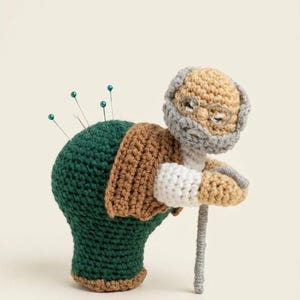 May include: A handcrafted crochet pincushion in the form of an elderly person, featuring a green body, brown jacket, and grey hair and beard. The pincushion has pins inserted into the back and holds a grey cane. The figure wears small, round spectacles.