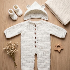 May include: A white crochet baby outfit set, featuring a hooded jumpsuit with brown buttons, matching booties, and a bear-eared hat. A wooden star teether and a folded blanket are also included.