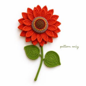Puede incluir: Un girasol de ganchillo con pétalos de color naranja vibrante, un centro marrón y dos hojas verdes en un tallo. El texto "pattern only" está escrito en letra verde a la derecha de la flor. El diseño presenta intrincadas puntadas de hilo.