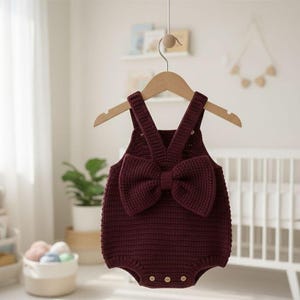 May include: A burgundy-colored baby romper with a large bow detail on the front. The knitted romper has suspender straps and wooden buttons. It is hanging on a wooden hanger.