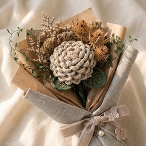 Pode incluir: Um buquê com uma flor central de crochê bege com folhas verdes, rodeada por plantas secas castanhas semelhantes a cardos e ramos verdes. O arranjo é embrulhado em papel castanho e tecido texturizado, atado com uma fita bege e detalhes de borboletas.