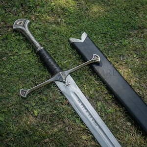 Puede incluir: Una espada plateada con un mango envuelto en cuero negro y un diseño de estrella en la hoja. La espada tiene una guarda y una vaina. La espada está sobre hierba verde.