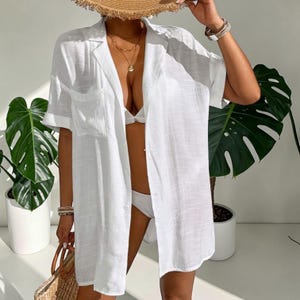 May include: A white, short-sleeved button-down shirt with a chest pocket, worn over a white bikini. A straw hat and a woven tote bag are also visible. The outfit is accessorized with gold necklaces and bracelets. Large green plants are in the background.