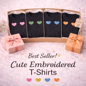 May include: Four black t-shirts with embroidered heart designs in pink, green, blue, and orange, presented in a cardboard box. The text "Best Seller! Cute Embroidered T-Shirts" is displayed below the shirts. Two gift boxes with bows are also present.