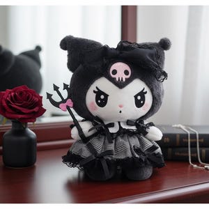 May include: A plush doll of a character with black fur, a skull on its head, and a plaid dress. The doll holds a trident with a pink bow. A red rose in a black vase and a stack of books are also visible.