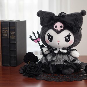 May include: A plush doll with a black and white checkered dress, black bow, and a skull on its head. It holds a trident with a pink bow. Black roses and books are in the background. The doll is approximately 8 inches tall.