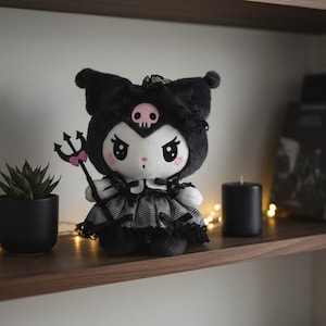 Könnte beinhalten: Schwarze Plüschpuppe mit Totenkopf auf dem Kopf und Dreizack. Die Puppe trägt ein schwarz-graues Kleid. Sie wird auf einem Holzregal mit einer kleinen Topfpflanze, einer schwarzen Kerze und Büchern ausgestellt.