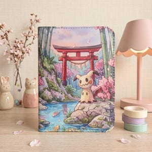 Könnte beinhalten: Ein rechteckiger Notizbuchumschlag mit einer farbenfrohen Illustration eines Mimikyu-Charakters, der auf einem Felsen an einem Bach steht, mit einem roten Torii-Tor, Bambusstangen und Kirschblütenbäumen im Hintergrund in sanften Pastelltönen.