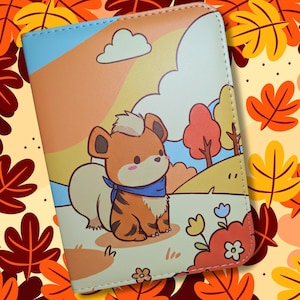 May include: A colorful wallet featuring a cartoon illustration of a brown and tan animal with a blue scarf, set against a landscape with trees and a rainbow. The background has a fall theme with orange and yellow leaves.