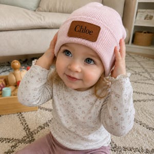 May include: A light pink ribbed knit beanie featuring a brown rectangular leather patch on the front with the name Chloe embossed in black text. The hat has a folded cuff design and is made of a soft, textured material suitable for headwear.
