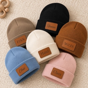 Puede incluir: Una colección de seis gorros de punto acanalado en negro, marrón, tostado, crema, azul y rosa. Cada gorro presenta un parche rectangular de cuero marrón centrado en el dobladillo, grabado con los nombres Rowan, Finn, Miles, Hazel, Elsie y Clara.