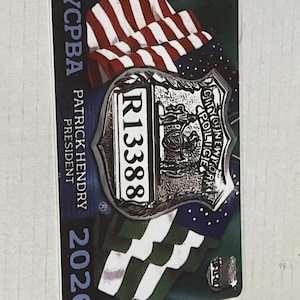 Könnte beinhalten: Eine rechteckige Karte mit einem silbernen Polizeiausweis mit dem Text "City of New York Police". Die Karte hat einen Hintergrund der amerikanischen Flagge und der Text "NYCPBA PATRICK HENDRY PRESIDENT 2026" ist auf die Karte gedruckt.