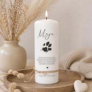 Pode incluir: Uma vela cilíndrica branca com pavio aceso, apresentando o nome Maja em escrita preta, uma pegada de pata preta e o texto em alemão PFOTEN HINTERLASSEN SPUREN FÜR DIE EWIGKEIT - NICHT AUF DEM BODEN, SONDERN IM HERZEN, com as datas 12.01.2020 e 01.06.2026.