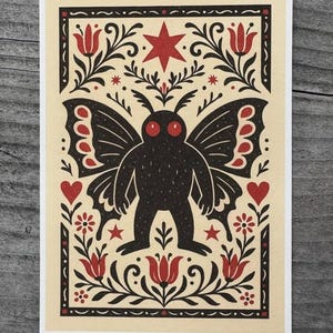 May include: A cream-colored art print featuring a black moth-like creature with red eyes and butterfly wings. The design includes red hearts, stars, and floral elements within a black border. The artwork has a folk art style.