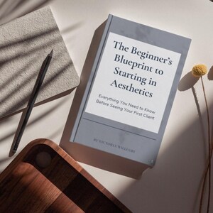 May include: A book titled "The Beginner's Blueprint to Starting in Aesthetics" with the subtitle "Everything You Need to Know Before Seeing Your First Client." A notebook, pencil, wooden tray, and dried flowers are also present.