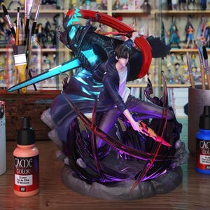 May include: A detailed figurine of a character in action, wielding a glowing blue sword and a smaller weapon. The figure is predominantly black and blue, with red accents. Paint bottles and brushes are visible in the foreground, suggesting a hobby or collectible item.