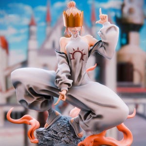 May include: A figurine of a person in a white outfit with a gold crown and a sun symbol. The figure is posed in a squatting position on a rock, with orange tendrils extending from the base. The background features a blurred cityscape with blue skies.