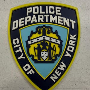 Puede incluir: Un emblema en forma de escudo con el texto "POLICE DEPARTMENT CITY OF NEW YORK" en letras blancas. El emblema presenta un diseño azul y amarillo con un escudo central y dos figuras. El diseño general es una insignia.