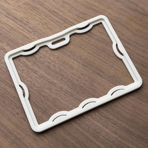 May include: A rectangular, off-white plastic badge holder frame featuring a scalloped inner edge design. The frame includes a centered horizontal slot at the top for attachment purposes, providing a clean, structured border for an identification card.