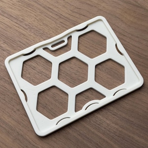 May include: A rectangular white plastic badge holder featuring a geometric honeycomb pattern with six hexagonal cutouts and a horizontal slot at the top for a lanyard attachment. The frame has rounded corners and a smooth, matte finish.