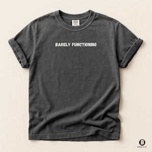 Puede incluir: Camiseta gris oscuro con mangas enrolladas. El texto blanco "BARELY FUNCTIONING" está impreso en el pecho. La camiseta tiene cuello redondo y una etiqueta que dice "COMFORT COLORS". El logotipo de la marca "BRICKLE CO." es visible en la parte inferior derecha.