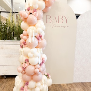May include: A balloon arch in shades of pink and white, adorned with small pink roses and white ribbons. The arch is set against a cream-colored backdrop with the text "BABY Penelope" in a delicate script, perfect for a baby shower.