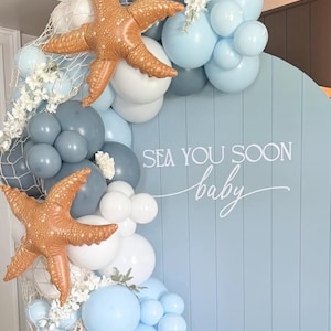 May include: A baby shower backdrop featuring a light blue wooden panel with the text "Sea You Soon baby". The backdrop is decorated with blue, white, and gray balloons, starfish, and white floral accents.
