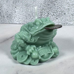 May include: A light green candle shaped like a Chinese money frog, holding a coin in its mouth. The frog sits on a pile of coins and has a banner with Chinese characters. The candle has a wick and is on a white surface.