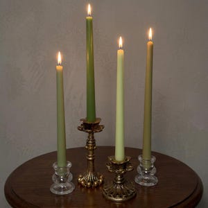 May include: Four lit taper candles in varying shades of green stand in four distinct candle holders. Two holders are made of clear glass with a tiered base, while the other two are crafted from ornate, textured gold-toned metal.