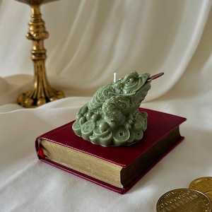 May include: A sage green wax candle shaped like a three-legged toad sitting on a pile of Chinese coins. The toad has a small metal coin in its mouth and a white wick protruding from its back. It is resting on a small red book with gold edges.