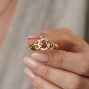 May include: A gold ring featuring three interlocking circular links. The central circular link is embellished with small, clear, sparkling stones set along its entire circumference, while the two outer links are smooth, polished gold.