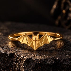 May include: A gold-toned ring featuring a geometric bat motif centered on a thin, polished band. The bat design displays sharp, angular wings and a pointed ear silhouette, crafted with a smooth, reflective metallic finish throughout the piece.