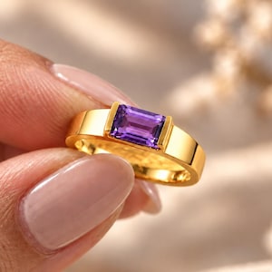Op de afbeelding: Een gouden ring met een rechthoekige, smaragdgeslepen paarse amethist die horizontaal in een gladde, gepolijste gouden band is gezet. De zetting houdt de levendige violette steen stevig vast tussen twee verticale gouden staven voor een strak ontwerp.
