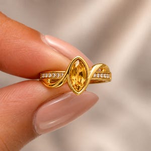 Può includere: Un anello in oro con una pietra preziosa gialla centrale taglio marquise in una montatura a lunetta. La fascia ha un design a bypass con una finitura in oro lucido ed è accentata con piccole pietre chiare incastonate a pavé sui lati.