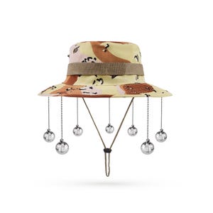 May include: A beige and brown camouflage bucket hat with a brown strap and a chin strap. The hat has silver disco ball-shaped ornaments hanging from the brim.