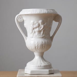 Może przedstawiać: Biała ceramiczna urna z dwoma bocznymi uchwytami i wypukłym wzorem przedstawiającym cherubiny z przodu. Naczynie posiada szeroki, rozszerzający się brzeg z dekoracyjnymi listwami, zwężający się korpus z pionowymi żłobieniami oraz podstawę na cokole.