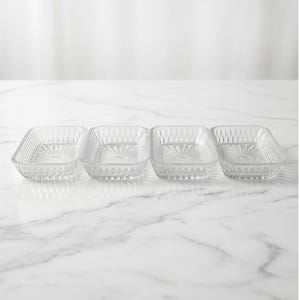 Peut inclure: Quatre plats rectangulaires en verre transparent avec des motifs de losanges et d'éclats d'étoiles. Les plats sont disposés en rangée, chacun avec un angle légèrement différent, sur fond blanc.