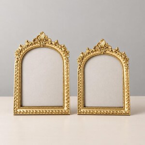 May include: Two arched picture frames with a metallic gold finish. Each frame features an ornate border with intricate floral and leaf detailing, a beaded inner edge, and a decorative crest at the top center of the arch.