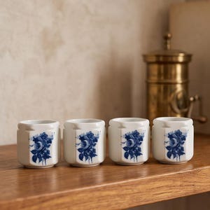 Peut inclure: Un ensemble de quatre tasses en céramique blanche, chacune ornée d'un motif floral bleu sur le devant. Les tasses présentent une forme cylindrique avec un bord légèrement facetté ou cranté. Le motif floral représente un bouquet de roses.